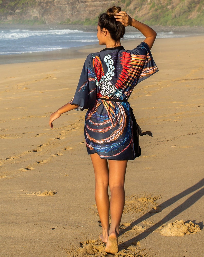 May include: A woman walks on a sandy beach wearing a black and blue kimono with a colorful phoenix design. The kimono has a black sash tied at the waist.