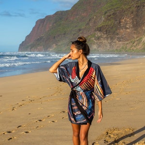 May include: A woman wearing a colorful patterned kimono-style dress walks along a sandy beach with a mountain range in the background.