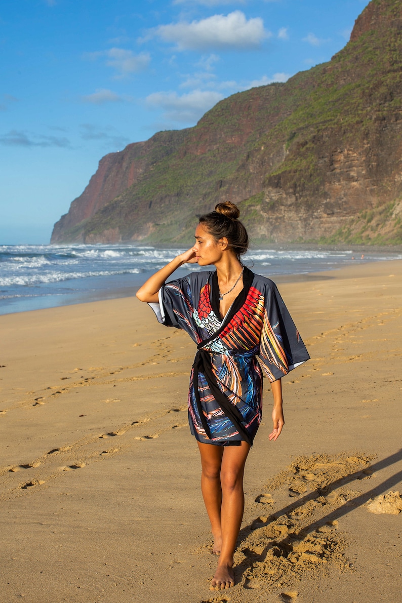 May include: A woman wearing a colorful patterned kimono-style dress walks along a sandy beach with a mountain range in the background.