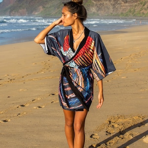 May include: A woman wearing a gray and black wrap dress with a colorful bird print. The dress has a black sash and a V-neckline. She is walking on a sandy beach.