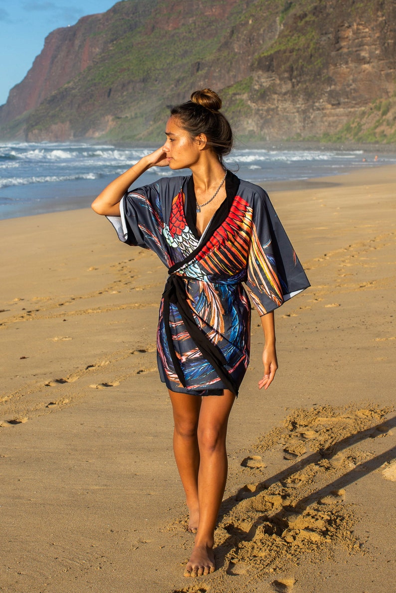 May include: A woman wearing a gray and black wrap dress with a colorful bird print. The dress has a black sash and a V-neckline. She is walking on a sandy beach.