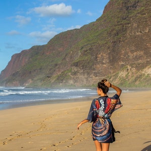 May include: A woman wearing a colorful patterned dress walks along a sandy beach with a mountain range in the background.