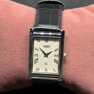 Puede incluir: Un reloj Seiko Quartz rectangular con esfera blanca, números romanos negros y correa de cuero negro. El reloj tiene una caja plateada y un pequeño dial de segundos. El reloj se muestra sobre una superficie rosa.