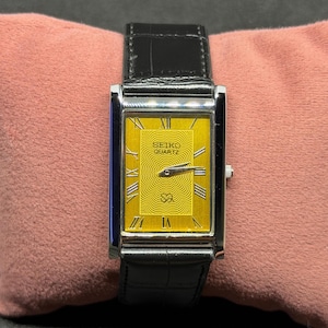 Pode incluir: Um relógio Seiko Quartz retangular com mostrador dourado e pulseira de couro preta. O mostrador do relógio apresenta algarismos romanos e o logotipo Seiko Quartz. O relógio é exibido em uma superfície rosa.