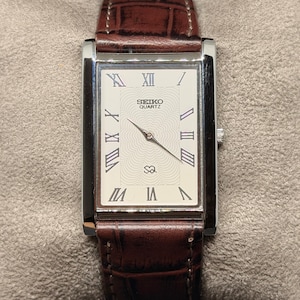 May include: Rectangular Seiko Quartz watch with a silver case and a brown leather band. The watch face is white with black Roman numerals and hands. The band has a crocodile pattern.