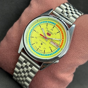 Pode incluir: Um relógio de pulso automático Seiko vintage com pulseira prateada e mostrador amarelo. O mostrador do relógio apresenta um ponteiro de segundos vermelho, um visor de dia e detalhes azuis e laranja. O relógio é exibido em uma superfície rosa.
