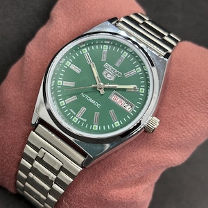 Peut inclure: Une montre automatique Seiko 5 argentée avec un cadran vert. La montre est dotée d'un bracelet argenté et d'un affichage de la date indiquant "DIM 27". La montre est présentée sur une surface rose.