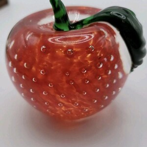 May include: A decorative glass apple with a red body and a green stem and leaf. The apple has small air bubbles throughout the red glass, and a white area on the side. The stem and leaf are a dark green color.