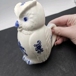 May include: A ceramic owl-shaped creamer with a white base and blue floral and geometric designs. The owl has blue eyes and a blue beak. The creamer has a handle and is held by a hand.