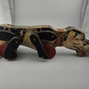 Op de afbeelding: Vintage houten trekdier in de vorm van een hond. De hond heeft een gevlekt lichaam, zwarte poten en rode wielen. De kop van de hond is beschilderd met een lachende uitdrukking en een rode halsband met "Snoopy". Een klassiek speelgoed.