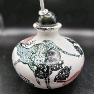 May include: A ceramic oil lamp with a white base, featuring a dark green and black abstract design. The lamp has a dark green top with a white wick. The lamp is a decorative item.