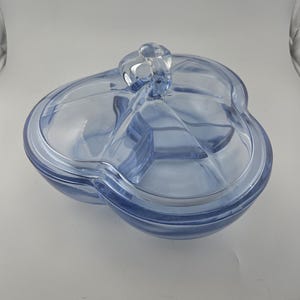 May include: A light blue, three-sectioned serving dish with a clear handle on the lid. The dish has a clover-like shape and is made of glass. The lid fits snugly over the sections, and the handle is looped for easy lifting.