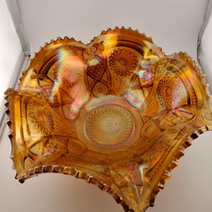 Puede incluir: Un cuenco de cristal de carnaval naranja iridiscente con un borde ondulado. El cuenco presenta un patrón detallado de diseños florales y geométricos. El cristal tiene una superficie brillante y reflectante, con toques de púrpura y verde a la luz.