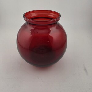May include: A round, red glass vase with a wide opening. The vase has a smooth, glossy surface and a deep red color. The vase is a decorative item that can be used to hold flowers or other items.