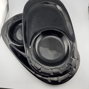 May include: Two black oval-shaped serving platters. Each platter has a central circular indentation and a raised, textured rim. The glossy finish reflects light, highlighting the smooth curves and contours of the serving dishes.