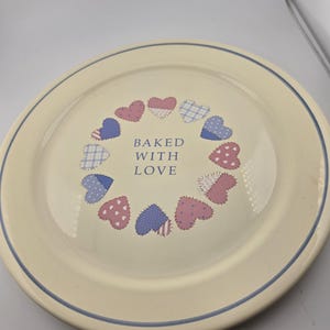 May include: A cream-colored ceramic plate with a blue rim and a ring of heart-shaped designs in red, blue, and white. The words "BAKED WITH LOVE" are in the center of the plate.