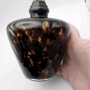 May include: A brown and black glass oil lamp with a decorative pattern. The lamp has a black top and a metal wick holder. The lamp is held by a hand.