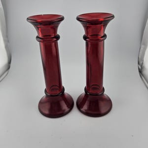 Puede incluir: Dos candelabros de vidrio rojo a juego. Cada uno tiene una base redonda, un tallo cilíndrico alto con un anillo decorativo y una parte superior acampanada. El vidrio es de un color rojo intenso y el diseño es simple y elegante.