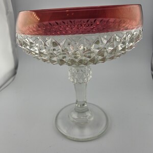 May include: A vintage champagne coupe glass with a ruby red rim and a clear, diamond-cut bowl. The glass has a delicate stem and a round base, perfect for serving celebratory drinks. The glass is approximately 6 inches tall.