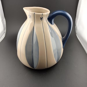 May include: A ceramic pitcher with a rounded body and a curved handle. The pitcher features vertical stripes in cream and blue, with small floral accents near the spout. The handle is a solid dark blue.