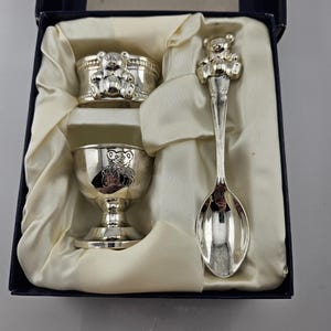 Peut inclure: Un coffret cadeau pour bébé en argent, comprenant un rond de serviette, un coquetier et une cuillère, tous ornés de motifs d'ours en peluche. L'ensemble est présenté dans une boîte bleue avec une doublure en satin, pour une présentation classique.