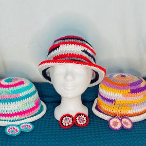 Può includere: Cinque cappelli all'uncinetto fatti a mano, colorati e con vari disegni, esposti su teste di manichino bianche. Ogni cappello presenta un motivo unico con tonalità vivaci. Orecchini all'uncinetto sono posti sotto ogni testa. I cappelli sono esposti su una superficie turchese.
