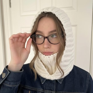 May include: Cream-colored knitted balaclava with a ribbed cowl neck and hood. The balaclava is made of a thick, textured yarn. The person in the image is wearing glasses and a denim jacket.