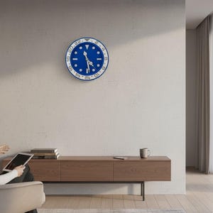 Puede incluir: Un reloj de pared azul y blanco con esfera redonda, que recuerda el diseño de un reloj, cuelga de una pared clara. El reloj tiene una esfera azul con marcas y manecillas blancas. La habitación cuenta con un mueble de madera, una ventana y una persona sentada.