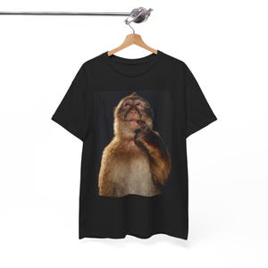 Peut inclure: T-shirt noir avec une image en gros plan d'un singe. Le singe a des poils bruns et se touche le menton avec sa main droite. Le t-shirt est suspendu à un cintre en bois. L'arrière-plan est noir.