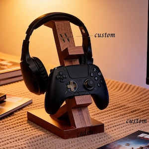 May include: A wooden stand for a gaming headset and controller. The stand is made of dark wood and holds a black headset and controller. The word "custom" is written on the stand. The stand is on a table with books.