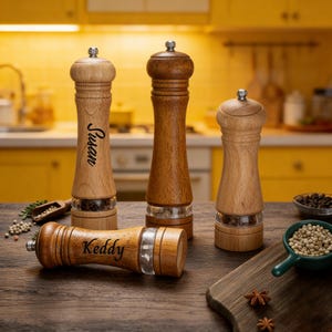 May include: A collection of wooden salt and pepper grinders in shades of brown, with clear glass bases. One grinder is lying down, displaying the name "Keddy." Two others have the name "Susan" on them. The grinders are on a wooden surface.