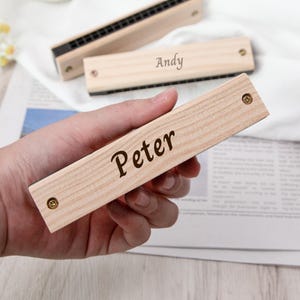 May include: Wooden harmonicas with black reeds and brass screws. One harmonica is held in a hand, with the name "Peter" engraved on it. Another harmonica has the name "Andy" engraved on it. The harmonicas are made of light-colored wood.