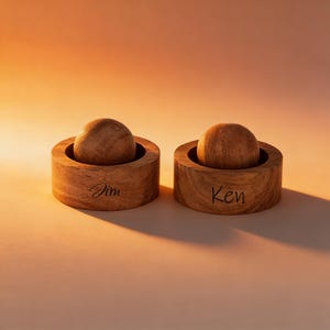 Może przedstawiać: Dwie drewniane, spersonalizowane solniczki i pieprzniczki. Każda z nich ma na górze okrągłą drewnianą kulkę, a na boku wygrawerowane imiona "Jim" i "Ken". Tło jest pomarańczowe.