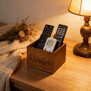Puede incluir: Un soporte de madera para mandos a distancia con la palabra "Remotes" grabada en la parte delantera. El soporte contiene tres mandos a distancia negros y un mando a distancia blanco. El soporte está sobre una mesa de madera junto a una lámpara.