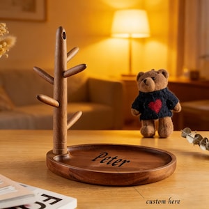 Op de afbeelding: Een houten mokkenboom met een ronde basis en een boomachtige structuur om mokken op te hangen. De basis heeft de naam "Peter" gegraveerd. Een kleine teddybeer met een trui met een rood hart staat op de achtergrond.
