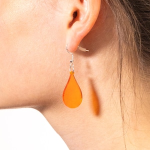 Può includere: Viene mostrato un singolo orecchino a goccia arancione. L'orecchino è sospeso a un gancio d'argento ed è realizzato in un materiale traslucido. L'orecchino è indossato sull'orecchio di una persona.