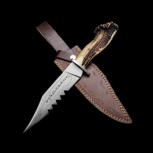 Może przedstawiać: Ręcznie wykonany nóż myśliwski z ząbkowanym ostrzem i rękojeścią z poroża jelenia. Nóż znajduje się w brązowej skórzanej pochwie z ozdobnymi przeszyciami. Ostrze ma wytrawiony tekst. Na czarnym tle.