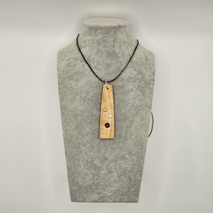 May include: A wooden pendant necklace with a rectangular, light-colored wood pendant. The pendant is adorned with three small, circular embellishments: gold, white, and red. The necklace cord is dark brown.