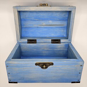 May include: Open blue wooden chest with a hinged lid and metal hardware. The chest has a rectangular shape with a distressed finish, showcasing the wood grain. The interior is also blue, and the chest is empty.
