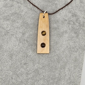 May include: A wooden pendant necklace with a brown cord. The pendant is rectangular with two circular insets, each containing a small, round, gold-colored bead. The pendant is made of light-colored wood.