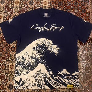 Pode incluir: T-shirt azul marinho com um gráfico de onda branca e o texto "Cough Syrup EST. 1988". O design lembra a arte japonesa. A camisa tem uma pequena etiqueta na manga e uma pequena etiqueta na parte inferior.