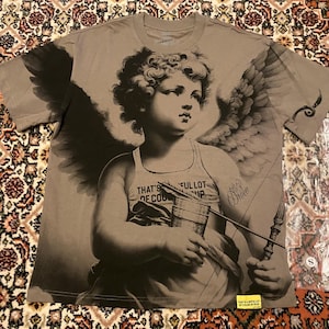 Könnte beinhalten: Taupefarbenes T-Shirt mit einem Graustufenbild eines Cherubs, der einen Bogen und einen Pfeil hält. Der Cherub trägt ein Tanktop mit Text. Das Shirt hat kurze Ärmel und einen Rundhalsausschnitt.