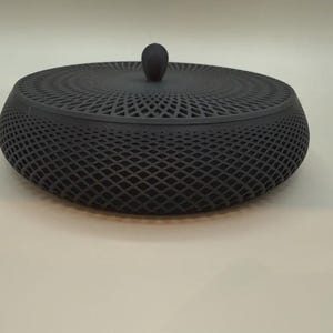 Pode incluir: Uma caixa decorativa preta e redonda com um design de treliça. A caixa tem uma tampa a condizer com um pequeno botão arredondado no topo. A caixa é feita de um material escuro e assenta num fundo neutro.