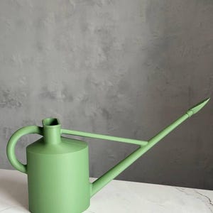 Puede incluir: Una regadera verde salvia con un pico largo y un asa curva. La regadera está hecha de un material sólido y se asienta sobre una superficie de mármol blanco. El fondo es una pared gris texturizada. Ideal para jardinería.