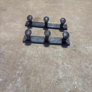 May include: Two sets of rustic, metal coat racks. Each rack has a dark, rectangular base with three rounded, textured knobs. The metal has a weathered appearance, suggesting a handmade or vintage origin. The racks are displayed on a concrete surface.