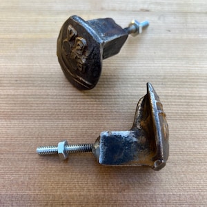 Könnte beinhalten: Zwei antike Schrankknöpfe aus Gusseisen mit einem dekorativen Emblem. Die Knöpfe sind dunkelbraun mit einem Used-Look-Finish und haben jeweils einen Gewindebolzen und eine Mutter zur Montage. Auf einer Holzoberfläche.