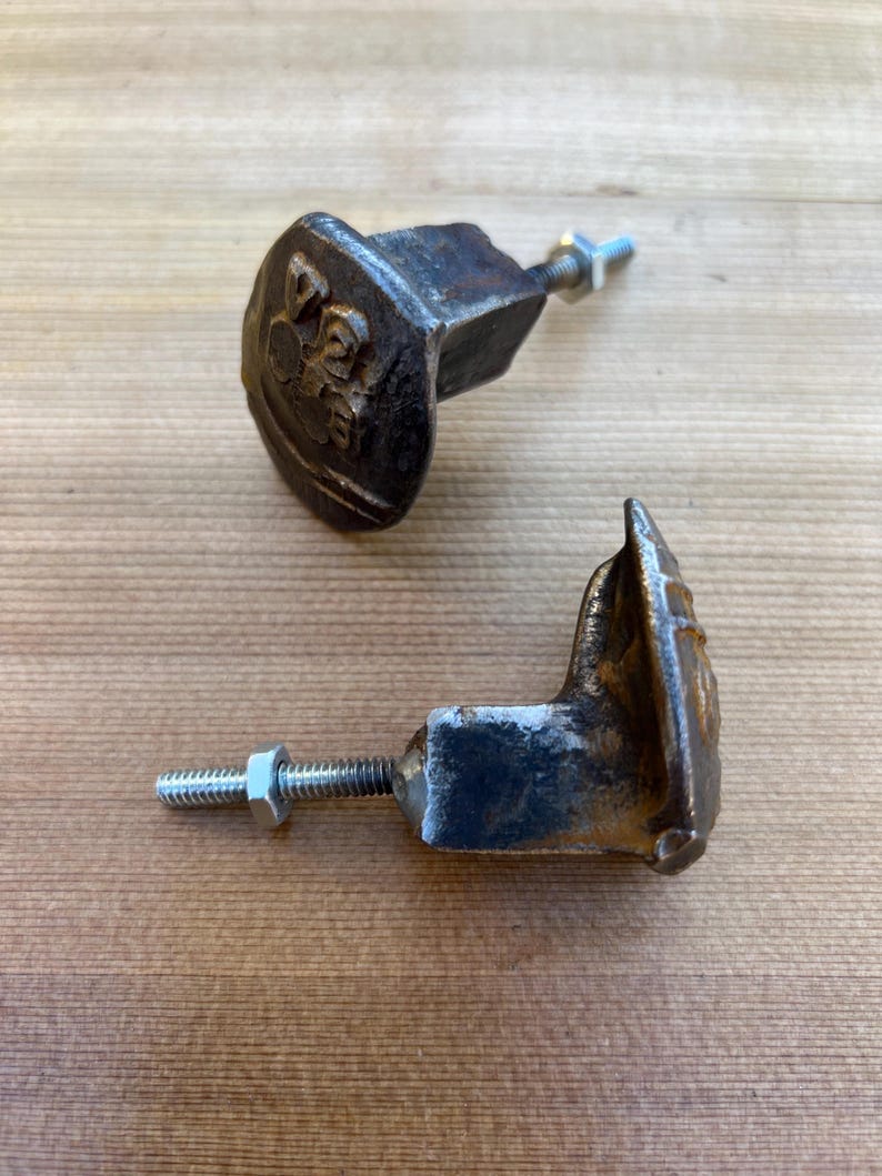 May include: Two antique, cast iron cabinet knobs with a decorative emblem. The knobs are dark brown with a distressed finish, and each has a threaded bolt and nut for mounting. The knobs are on a wooden surface.