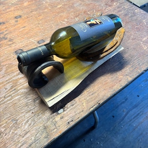 May include: A handcrafted wine bottle holder made from a horseshoe and wood. The holder supports a green wine bottle, displaying a unique combination of materials. The horseshoe is a rusty brown colour, and the wood has a natural finish.