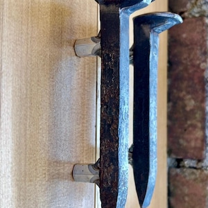 Könnte beinhalten: Zwei rustikale, dunkle Metalltürgriffe in Form von Bahnschwellennägeln. Die Griffe sind an einer hellen Holztür befestigt, im Hintergrund ist eine Backsteinmauer zu sehen. Das Metall hat ein verwittertes, gealtertes Aussehen.