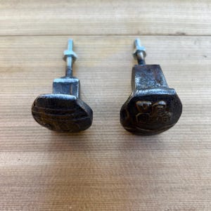 May include: Two weathered, dark metal cabinet knobs with an oval design. Each knob has a threaded bolt and nut for attachment. The knobs are set against a light brown wooden surface.